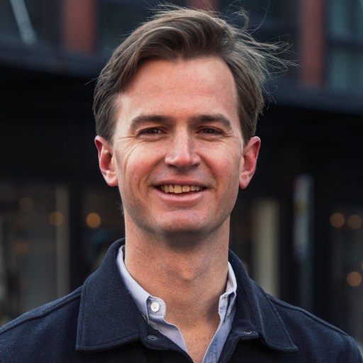 A man with light brown hair, wearing a dark jacket over a light shirt, stands outdoors and smiles at the camera.