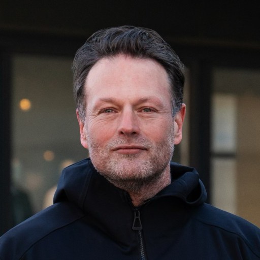 A middle-aged man with short dark hair and a trimmed beard, wearing a black jacket, stands outdoors in front of a blurred building background.