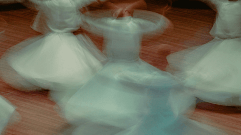 Blurred image of people in white robes spinning in a circular motion on a wooden floor, creating a sense of movement and flow.