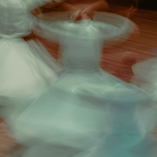 Blurred image of people in white robes spinning in a circular motion on a wooden floor, creating a sense of movement and flow.