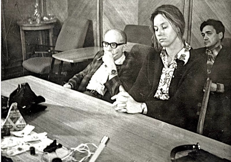 Three people sit at a table in a room; various items, including papers and small objects, are spread out on the table in front of them.