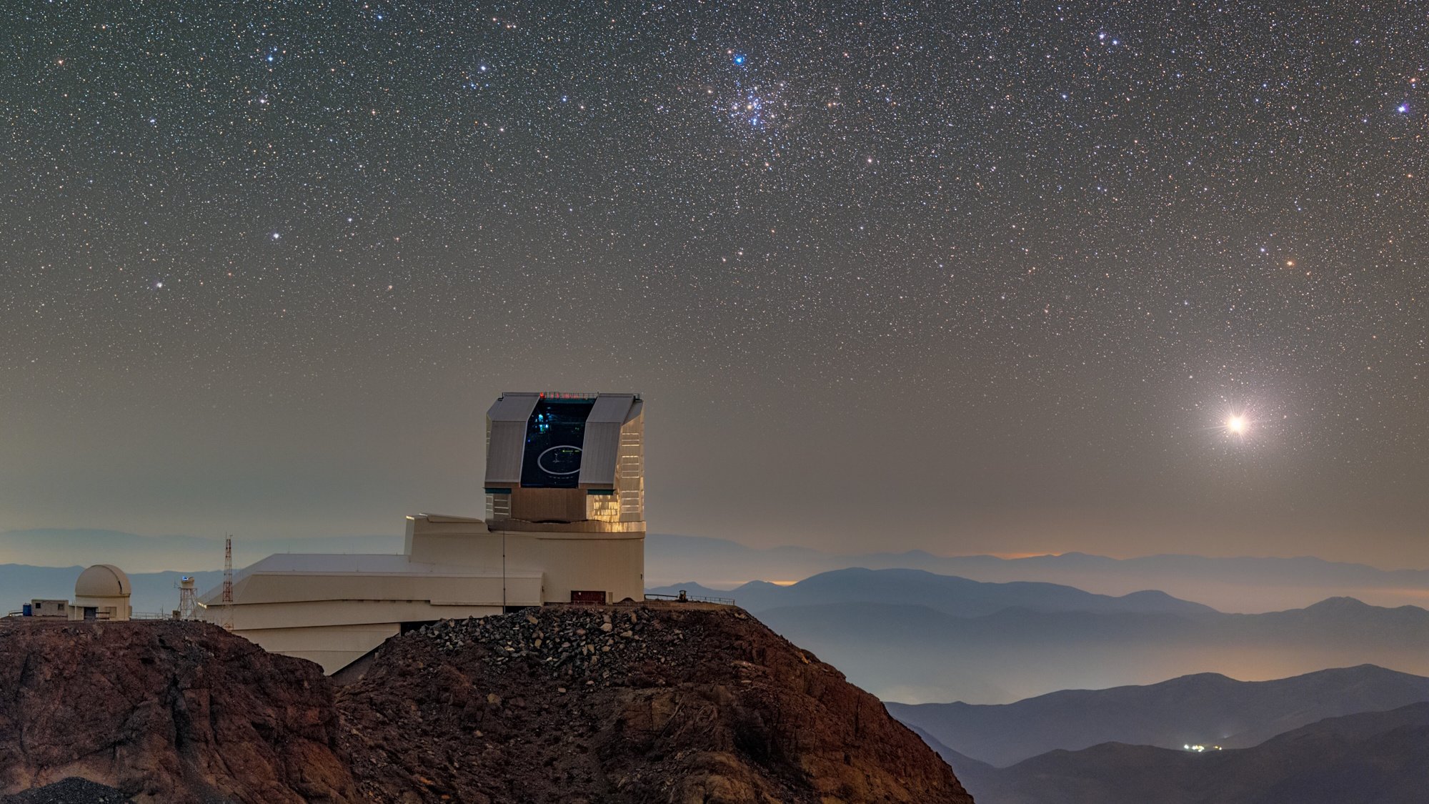 The Vera Rubin Observatory is situated on a rocky hilltop under a clear, star-filled night sky, with distant mountains and a bright planet visible on the horizon, inspiring astronomers to solve puzzles of the universe.
