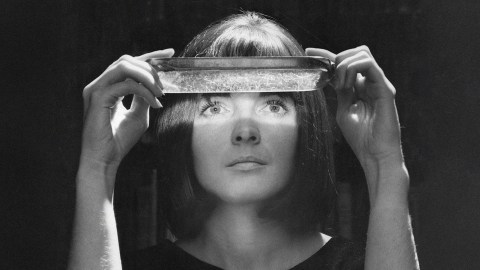 A woman, resembling a paranormal investigator, holds a rectangular glass dish above her eyes, which are illuminated by light shining through the dish in a dark setting.