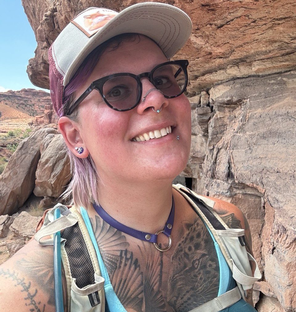 Person with glasses, a cap, and visible tattoos smiles while hiking in a rocky, desert-like outdoor area.