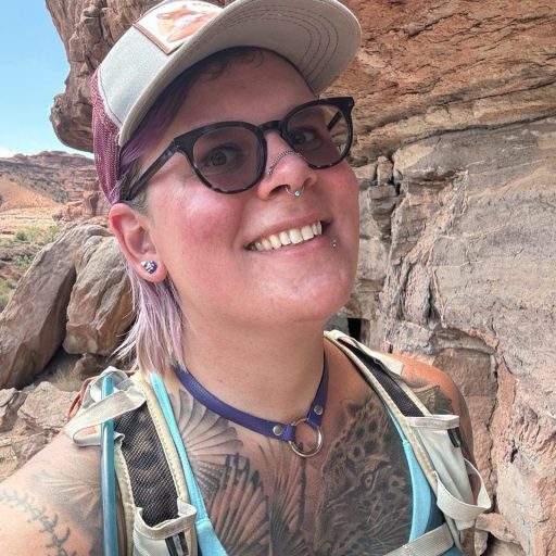 Person with glasses, a cap, and visible tattoos smiles while hiking in a rocky, desert-like outdoor area.