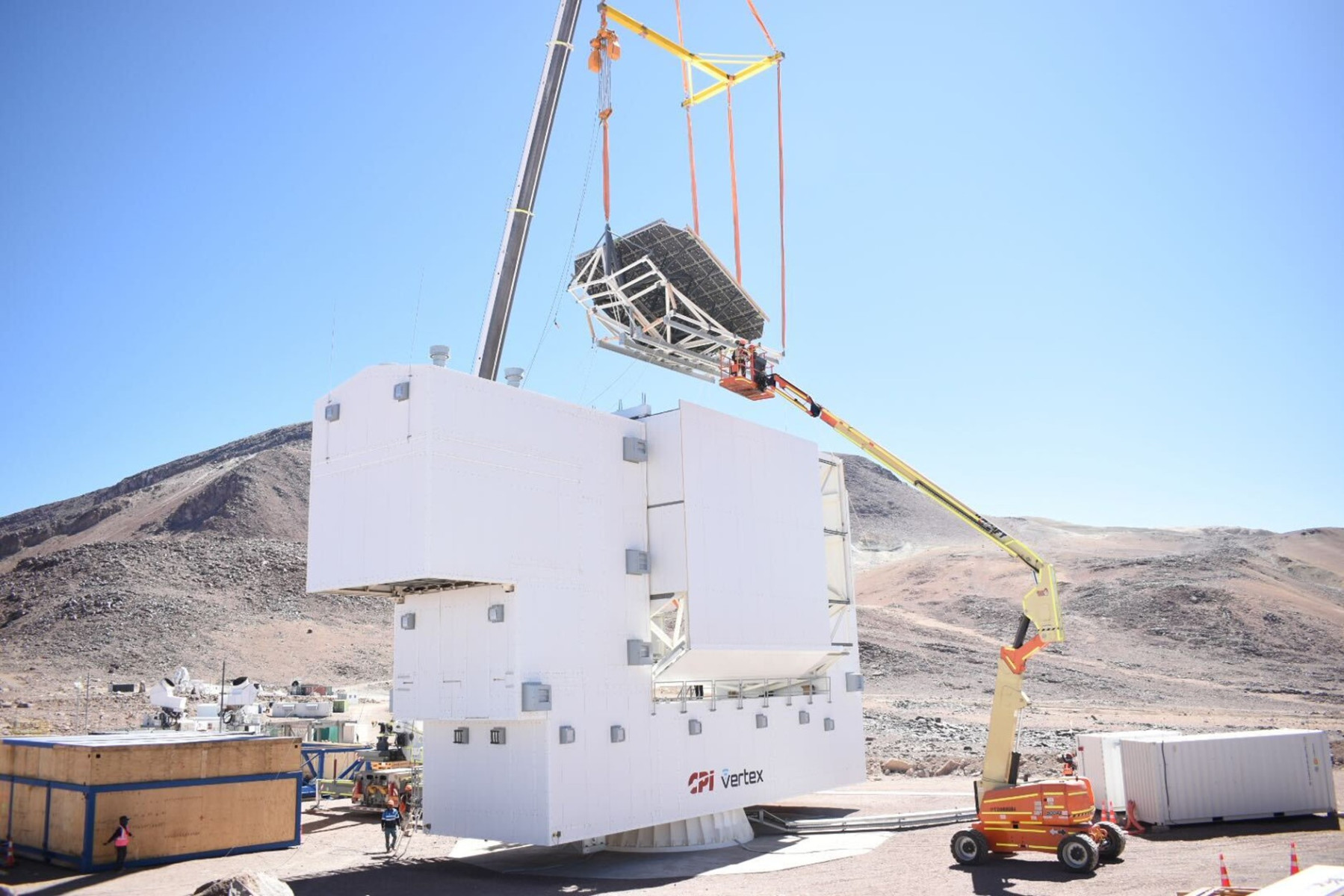 A crane lifts a large metal structure onto a white building at a construction site in a mountainous, arid area under clear blue sky.