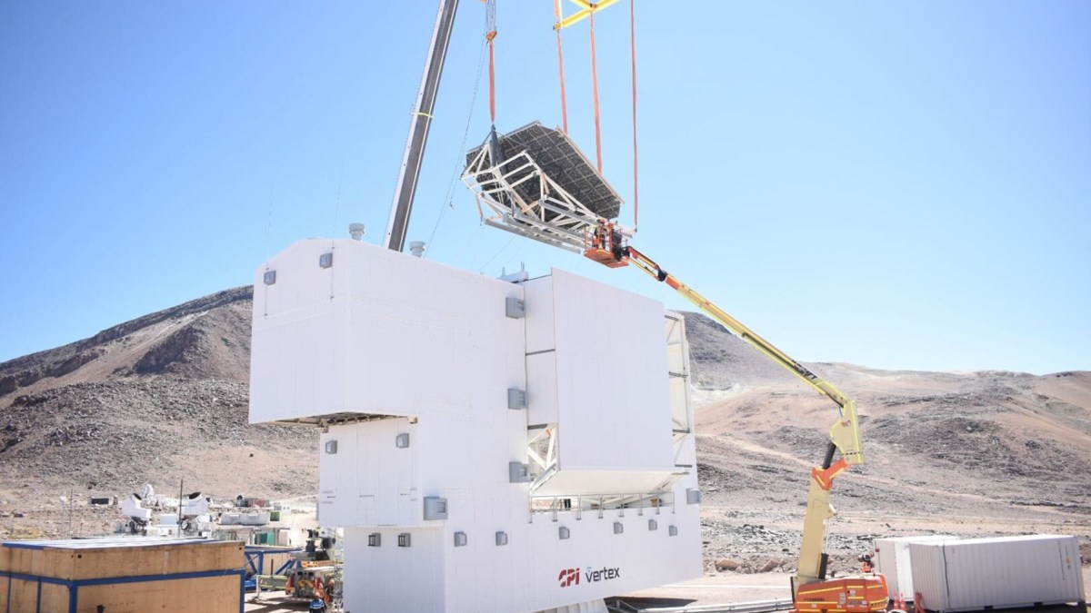 A crane lifts a large metal structure onto a white building at a construction site in a mountainous, arid area under clear blue sky.