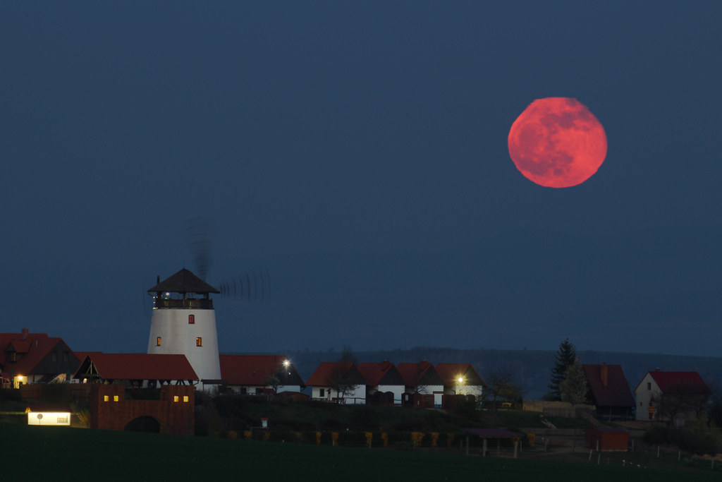 A large red moon rises over a village with a windmill and houses at dusk, some lights glowing in buildings—an inspiring scene perfect for 5 science lessons about the moon.