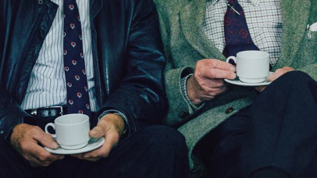 Two men in suits sit side by side, each holding a white cup and saucer, with only their torsos and hands visible—one could easily imagine Aaron Hurst sharing a thoughtful conversation over coffee.