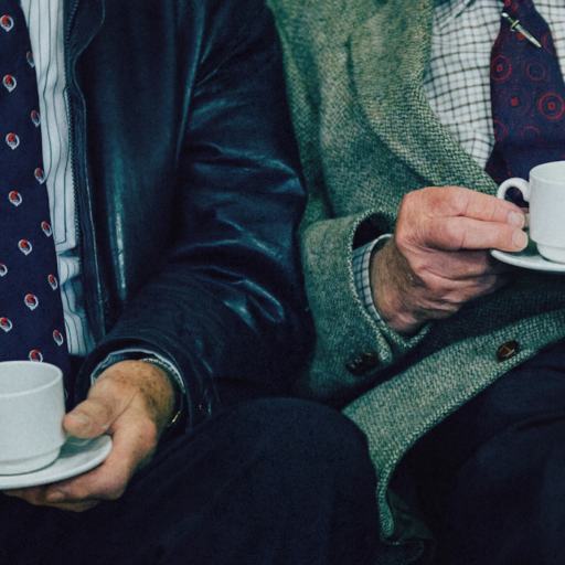 Two men in suits sit side by side, each holding a white cup and saucer, with only their torsos and hands visible—one could easily imagine Aaron Hurst sharing a thoughtful conversation over coffee.
