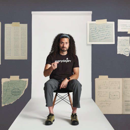 A man with long curly hair sits on a chair in a white hallway, surrounded by illustrated documents, maps, and notes taped to a gray wall.