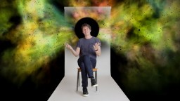A man sits on a stool in front of a white backdrop with a black circle behind his head, surrounded by colorful, nebula-like clouds.