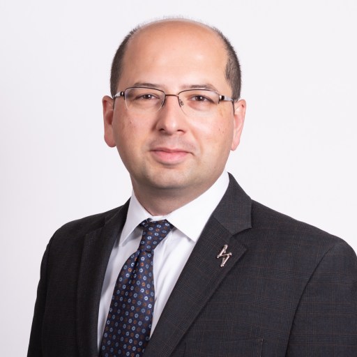 A man wearing glasses and a dark suit with a patterned tie poses against a plain white background.