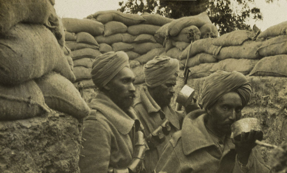Three soldiers wearing turbans sit in a sandbagged trench; one drinks from a metal cup while the others look ahead.