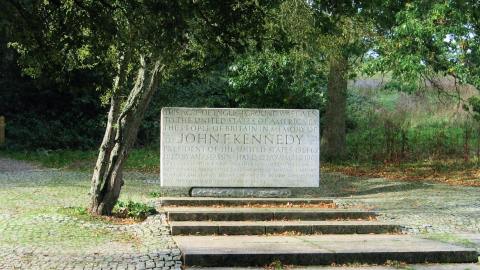Granite memorial stone for John F. Kennedy, surrounded by trees and located on a paved area with steps. Inscription dedicates the site from the people of Britain to the United States.