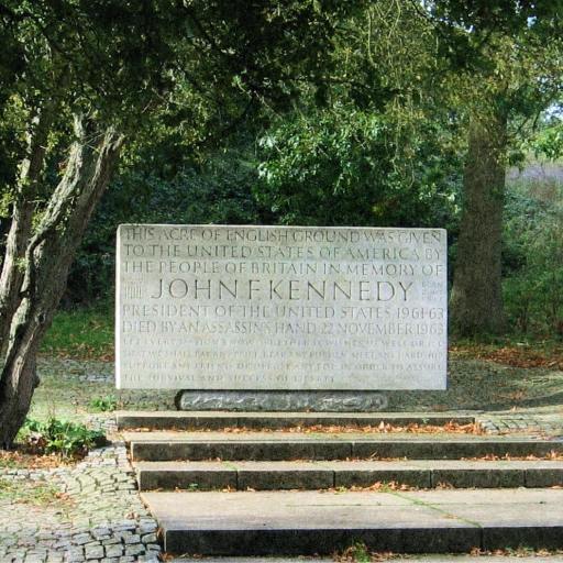 Granite memorial stone for John F. Kennedy, surrounded by trees and located on a paved area with steps. Inscription dedicates the site from the people of Britain to the United States.