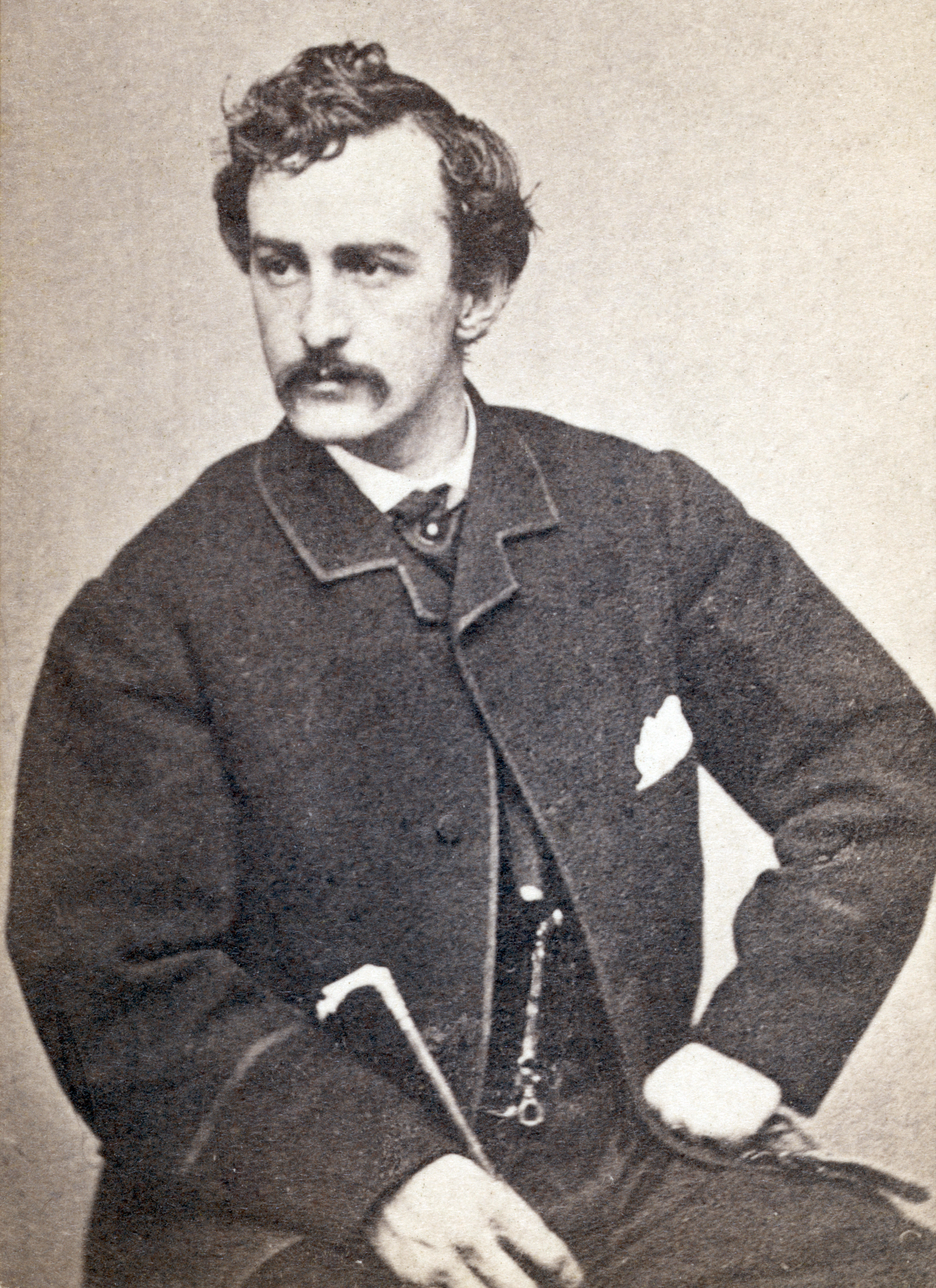 A man with dark hair and a mustache sits, wearing a suit with a pocket watch chain and holding a cane, facing slightly to the side.