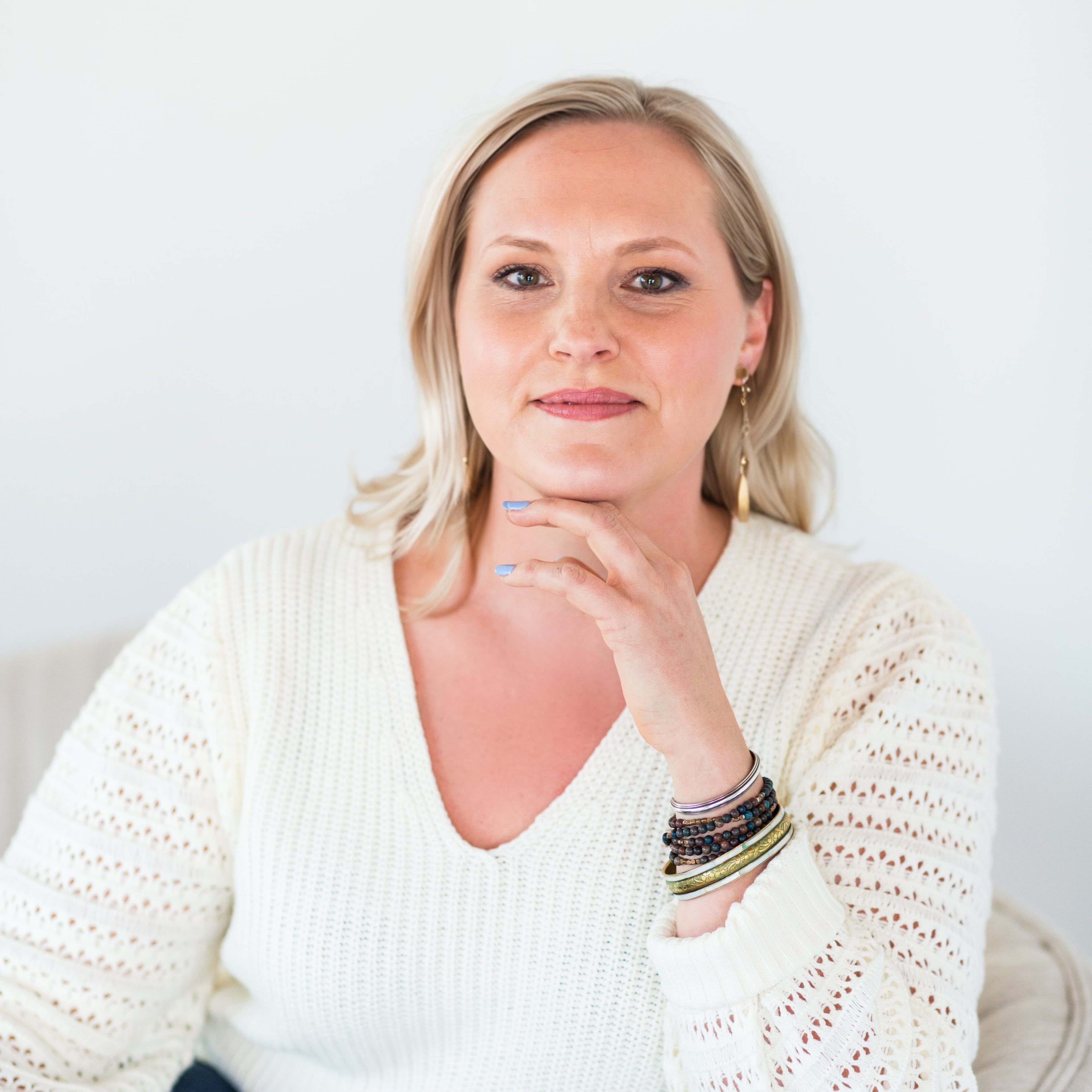 A woman with blonde hair sits in a light-colored chair, wearing a white sweater and jeans, resting her hand on her chin and looking at the camera.