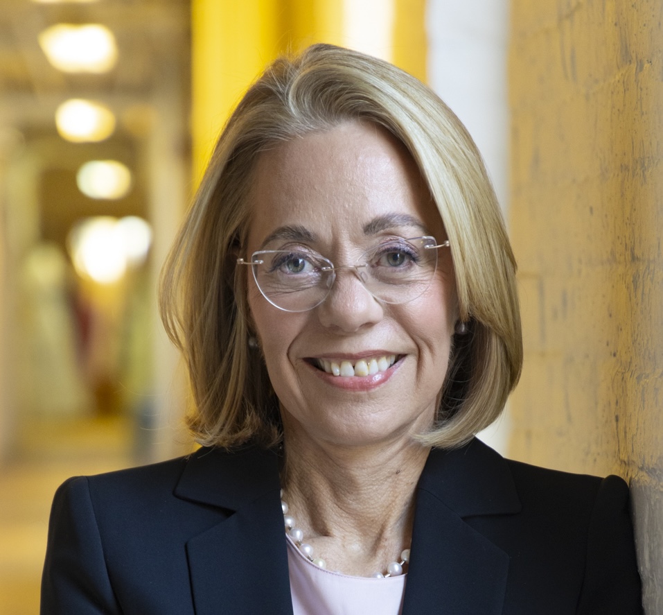 A woman with blonde hair and glasses, wearing a black blazer and a pearl necklace, stands indoors in front of a yellow wall, smiling at the camera.