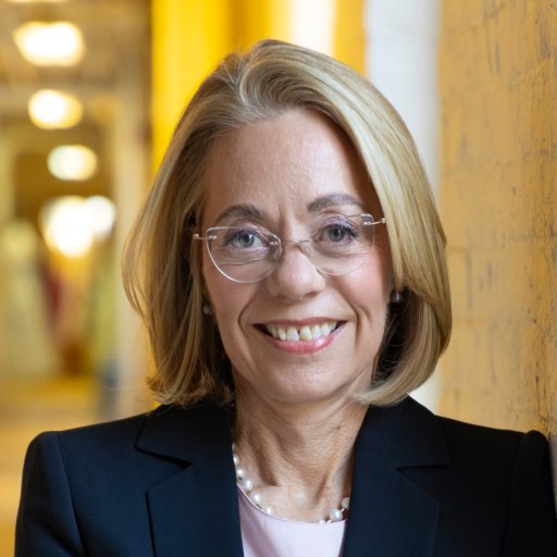 A woman with blonde hair and glasses, wearing a black blazer and a pearl necklace, stands indoors in front of a yellow wall, smiling at the camera.