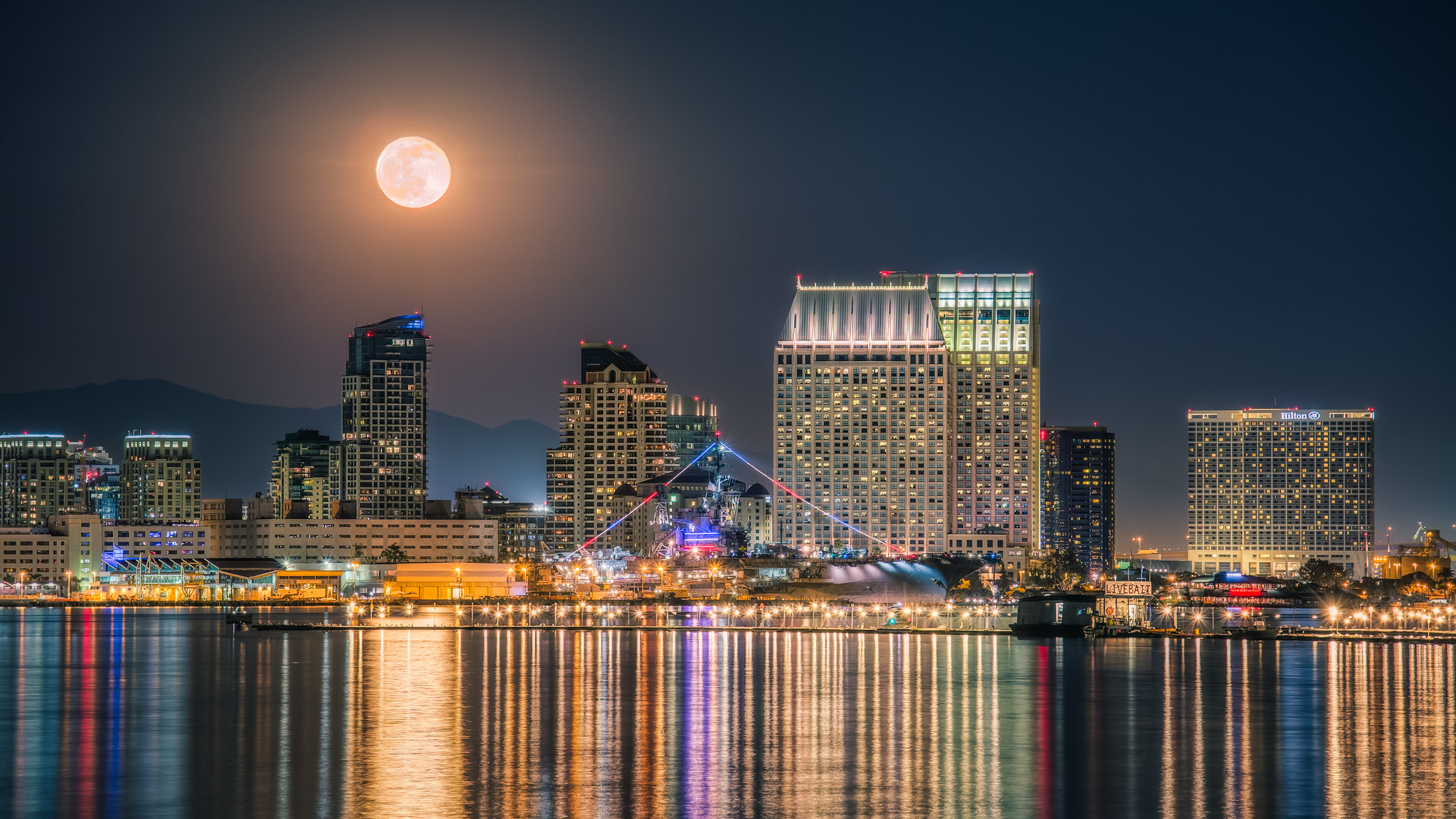 Full moon over a city skyline at night, high-rise buildings aglow and lights reflecting on the calm water—a scene that inspires 5 science lessons about the moon's impact on our world.