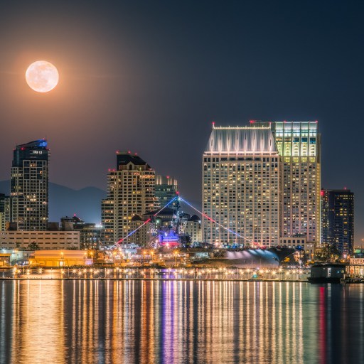 Full moon over a city skyline at night, high-rise buildings aglow and lights reflecting on the calm water—a scene that inspires 5 science lessons about the moon's impact on our world.