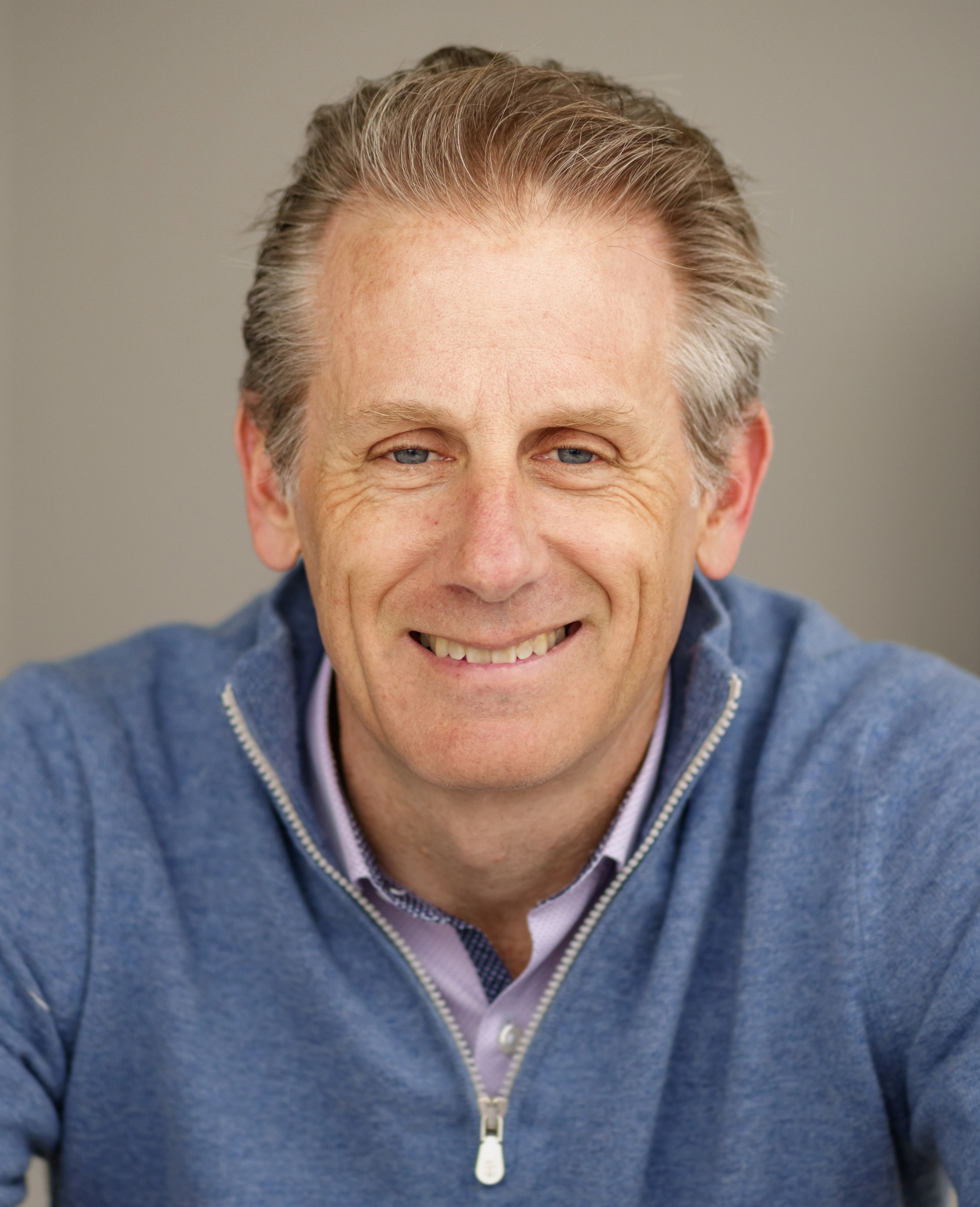 A middle-aged man with light brown hair wearing a blue zip-up sweater and a collared shirt, smiling at the camera against a neutral background.
