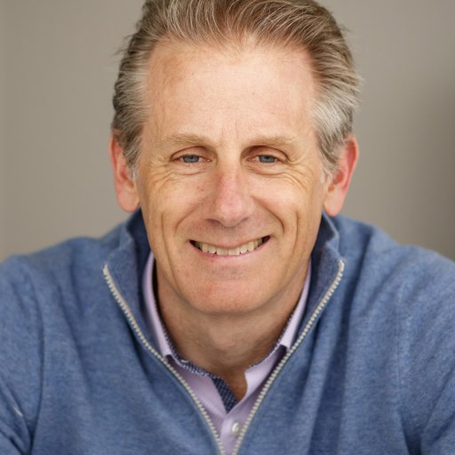 A middle-aged man with light brown hair wearing a blue zip-up sweater and a collared shirt, smiling at the camera against a neutral background.