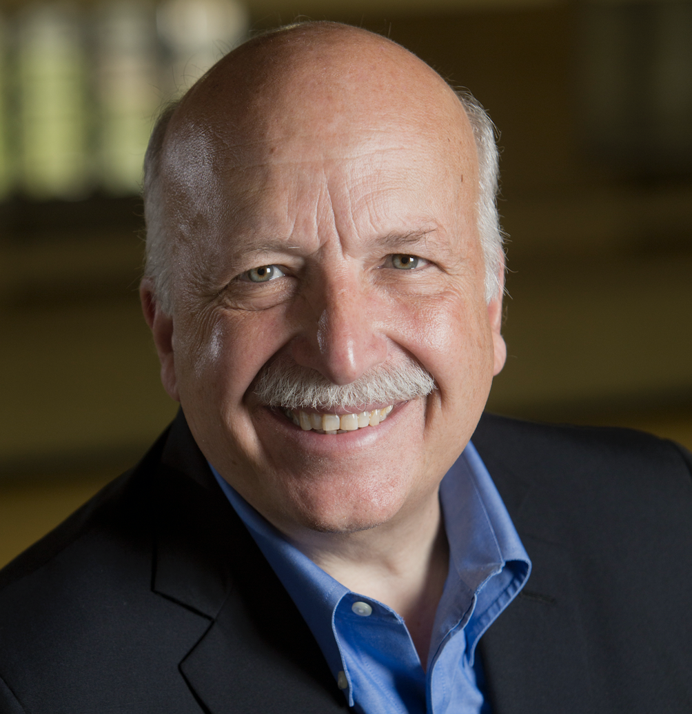 Smiling older man with a bald head, gray mustache, and blue shirt under a dark blazer, posing in front of a blurred indoor background.
