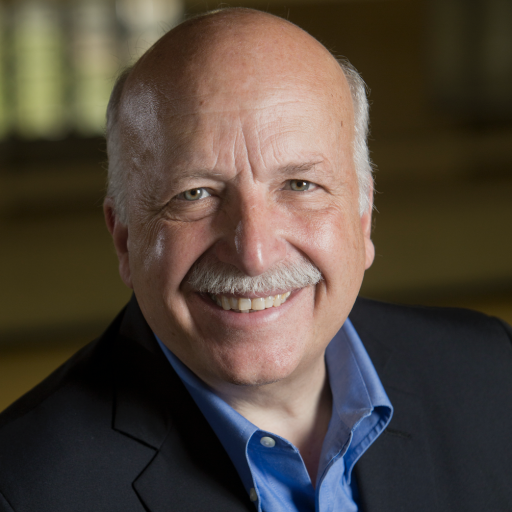 Smiling older man with a bald head, gray mustache, and blue shirt under a dark blazer, posing in front of a blurred indoor background.