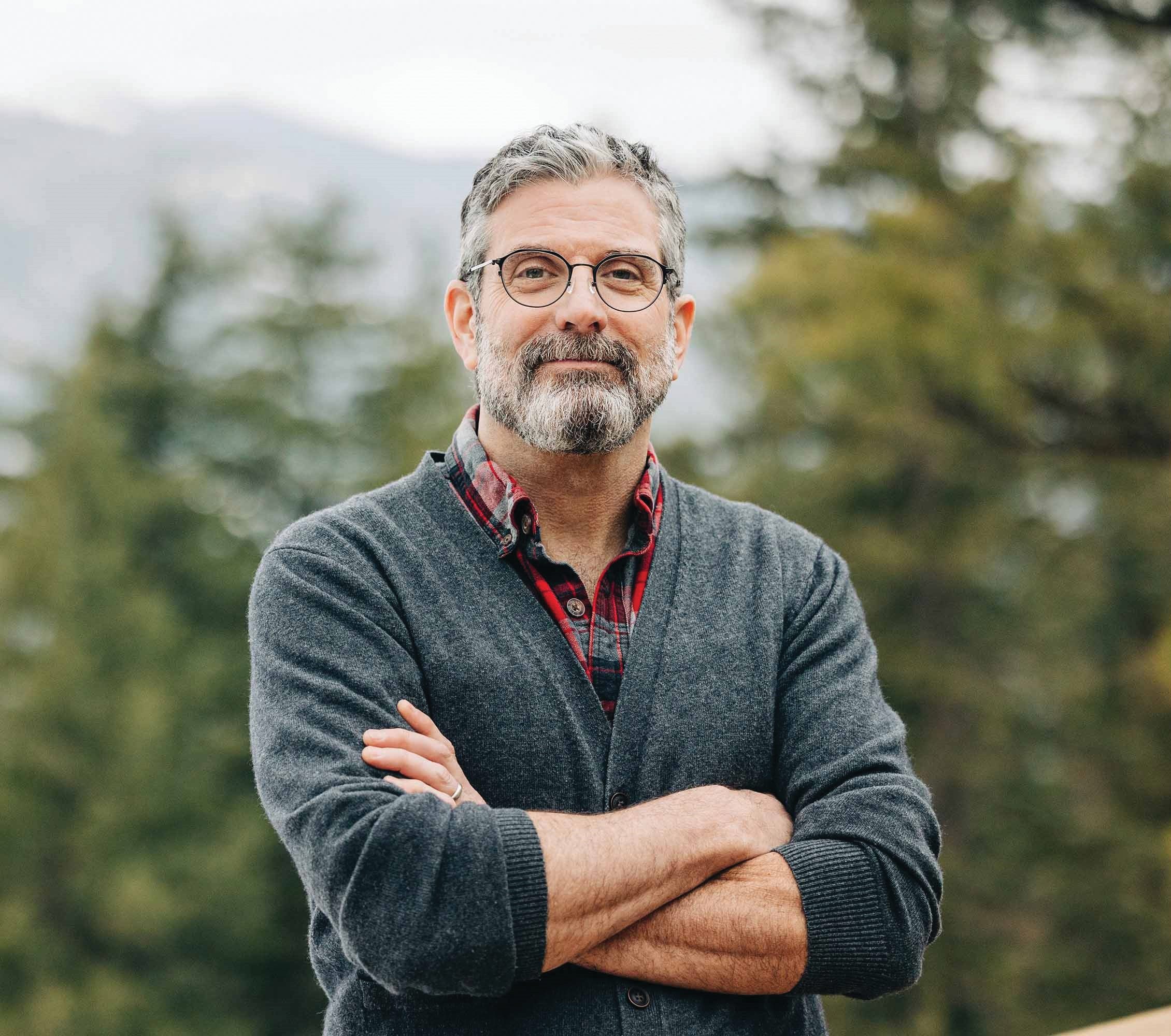 Man with gray hair and beard wearing glasses and a cardigan stands outdoors with arms crossed, trees and mountains in the background.