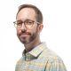 A man with short brown hair, beard, and glasses, wearing a yellow and blue plaid shirt, poses against a white background.