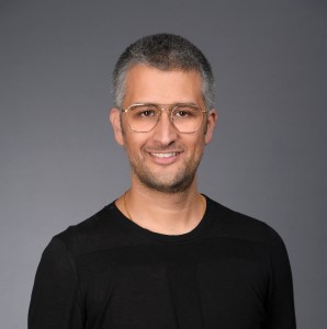 A man with short gray hair and glasses, wearing a black shirt, poses in front of a plain gray background, smiling slightly.
