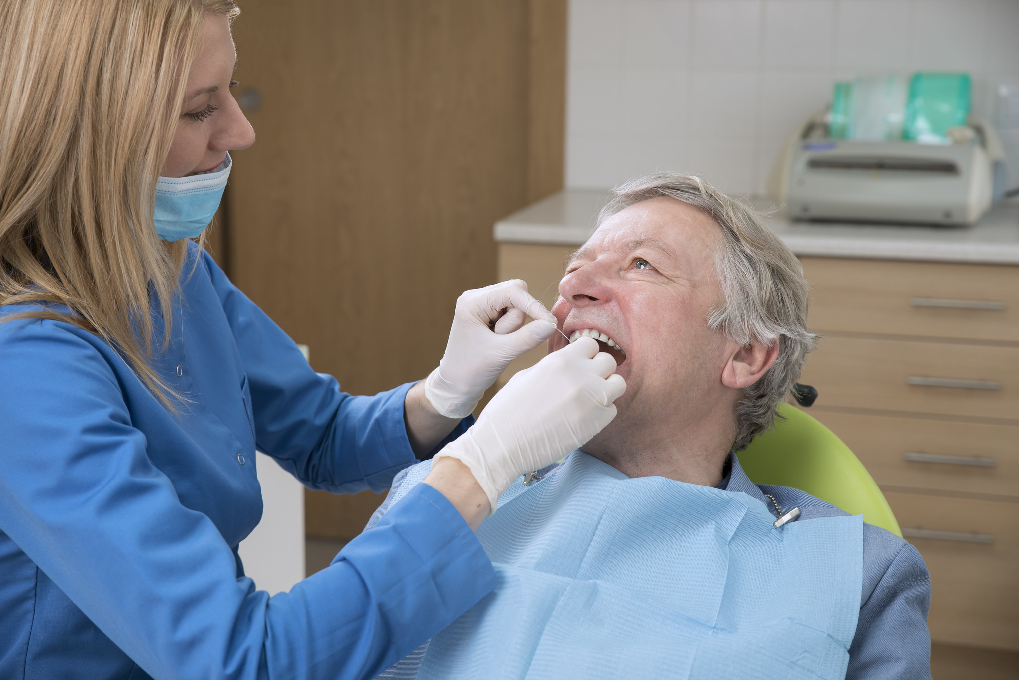 A dentist examines an older male patient's mouth while he sits in a dental chair wearing a blue bib, reminding us—like Einstein said—you can’t change the facts, but you can care for your smile.
