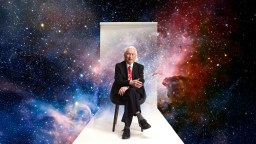 An older man in a suit and red tie sits on a chair against a white backdrop, with a colorful outer space scene in the background.