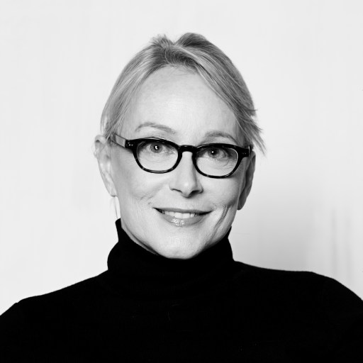 Woman with light hair wearing glasses and a black turtleneck, smiling at the camera against a plain background.