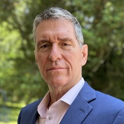 A middle-aged man with short gray hair wearing a blue blazer and light pink shirt, standing outdoors with trees in the background.