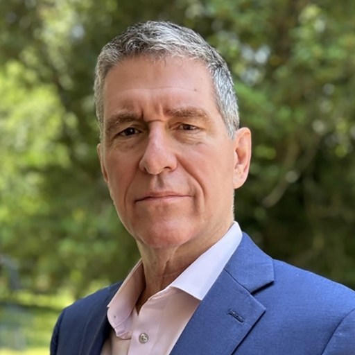 A middle-aged man with short gray hair wearing a blue blazer and light pink shirt, standing outdoors with trees in the background.