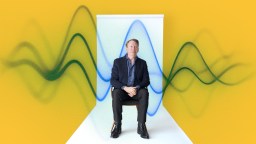 A man in a suit sits on a chair against a yellow background with abstract blue and green wave patterns behind him.