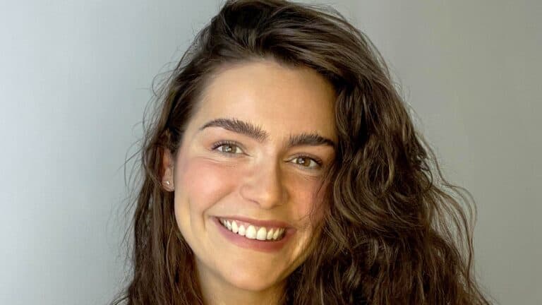 A woman with long wavy brown hair and thick eyebrows is smiling at the camera against a plain light gray background.