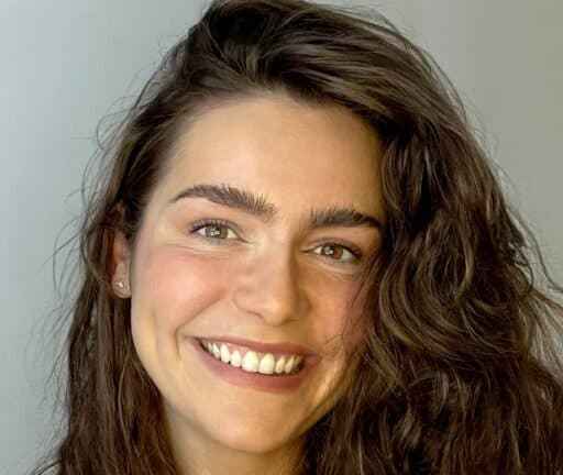 A woman with long wavy brown hair and thick eyebrows is smiling at the camera against a plain light gray background.