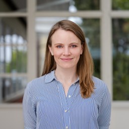 A woman with straight light brown hair, wearing a blue and white striped button-up shirt, stands indoors in front of large windows.