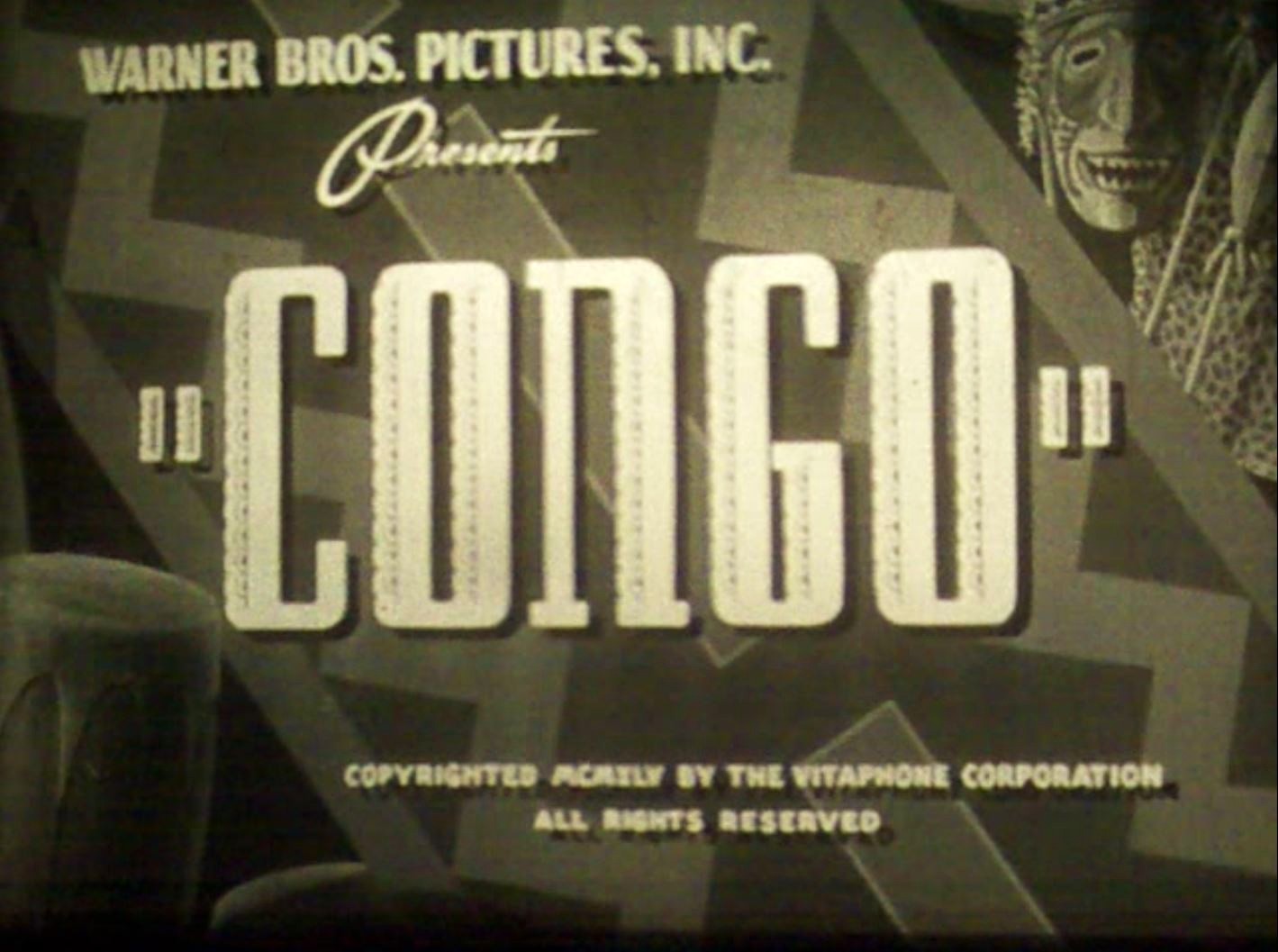 Black-and-white title card for the film "Congo," presented by Warner Bros. Pictures, with tribal masks and drums in the background.
