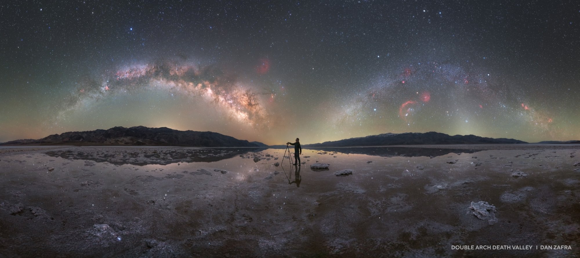 A person stands on a reflective salt flat under a starry night sky with the Milky Way arching overhead, capturing the essence of 2025 night sky images in Death Valley.