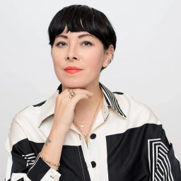 Ingrid Hu Dahl with short black hair and bangs, wearing a black and white patterned shirt, layered necklaces, and a ring, posing against a plain white background.