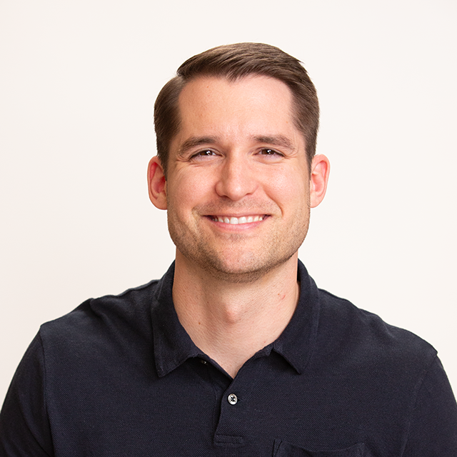 A man with short brown hair, wearing a dark polo shirt, smiles at the camera against a plain light background.