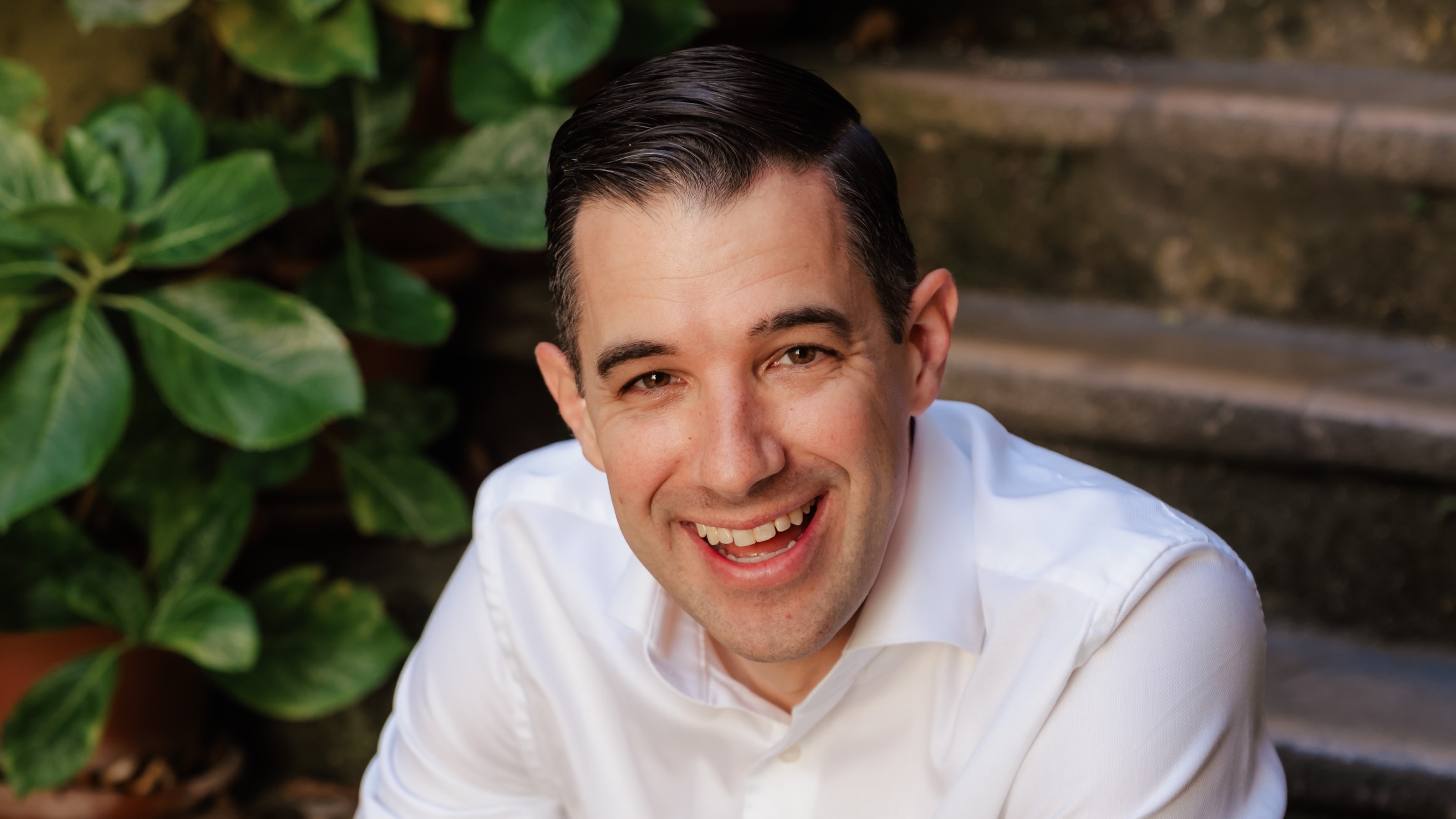 John Candeto, in a white shirt, smiles while sitting outdoors near stone steps and green potted plants.