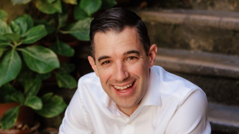 John Candeto, in a white shirt, smiles while sitting outdoors near stone steps and green potted plants.