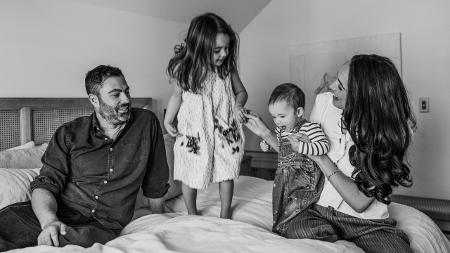 A man and woman sit on a bed with two young children, sharing a quiet moment; the older child stands while the younger, cradled by the woman, embodies the precious gift of consciousness within the family.
