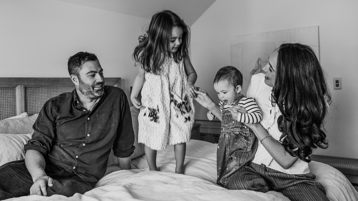 A man and woman sit on a bed with two young children, sharing a quiet moment; the older child stands while the younger, cradled by the woman, embodies the precious gift of consciousness within the family.